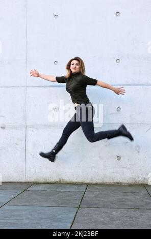 jeune femme en vêtements noirs sautant devant le mur gris Banque D'Images
