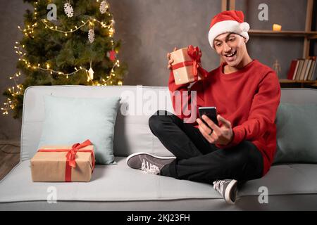 Jeune homme heureux blogueur dans le chapeau de santa montrant le présent sur l'appareil photo de smartphone assis sur le canapé à la maison le soir de noël. Positif gars se réjouissant à xmas g Banque D'Images