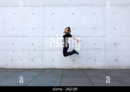 jeune femme en vêtements noirs sautant devant le mur gris Banque D'Images