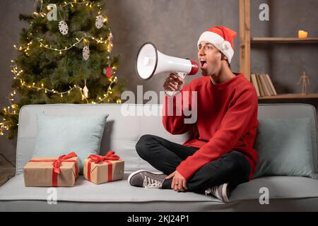 Un jeune homme heureux dans le chapeau du Père Noël criant en utilisant le mégaphone tout en étant assis à la maison. Partager des nouvelles sur les ventes saisonnières. Concept de vente de Noël Banque D'Images