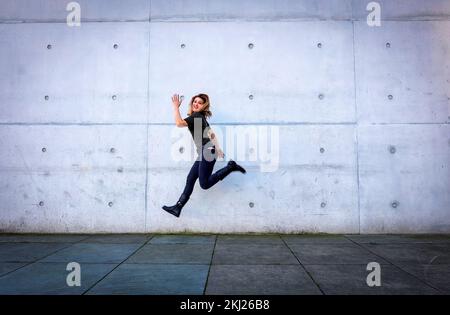 jeune femme en vêtements noirs sautant devant le mur gris Banque D'Images