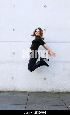 jeune femme en vêtements noirs sautant devant le mur gris Banque D'Images