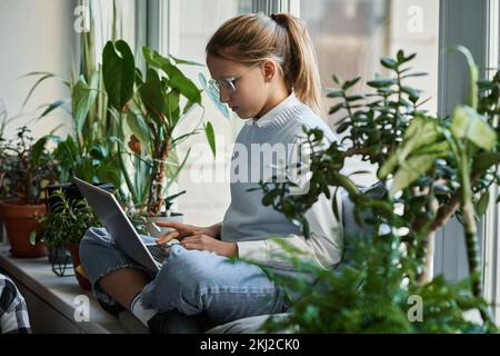 Écolière à lunettes se concentrant sur la communication en ligne, elle s'asseyait sur windowsill et tapant sur ordinateur portable parmi les plantes vertes Banque D'Images