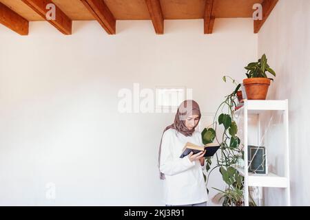 Jeune femme en foulard lire un livre debout près de la bibliothèque. Jeune fille musulmane se préparant aux examens d'entrée. Concept d'éducation Banque D'Images