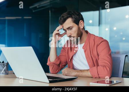 Triste et pensant programmeur en chemise rouge travaillant sur ordinateur portable à l'intérieur du bureau, homme mûr avec la barbe. Banque D'Images