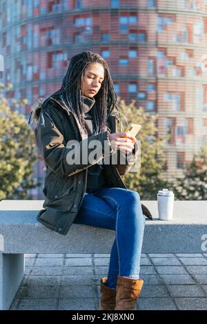 Femme hispanique concentrée dans les messages texte de vêtements d'extérieur sur téléphone portable tout en étant assis sur un banc de pierre avec thermos de café chaud dans la ville Banque D'Images