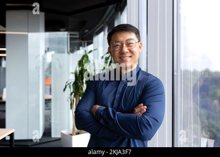 Portrait d'un homme d'affaires asiatique réussi, homme à l'intérieur du bureau portant une chemise et des lunettes souriant et regardant la caméra, courtier d'investisseurs avec les bras croisés près de la fenêtre. Banque D'Images
