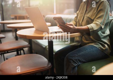 Gros plan de la main d'une femme utilisant un smartphone tout en travaillant sur un ordinateur portable dans un café. Banque D'Images