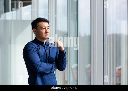 Homme d'affaires asiatique prospère pensant par la fenêtre, homme en chemise et lunettes investisseur pensant à des projets futurs. Banque D'Images