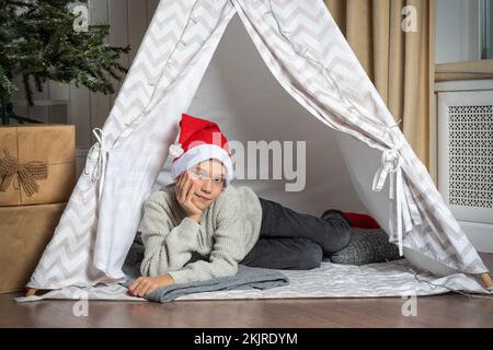 Un jeune garçon dans un chapeau de père Noël allongé dans une tente pour enfants dans une chambre qui attend impatiemment pour le père Noël. Le concept des vacances de Noël et du nouvel an Banque D'Images