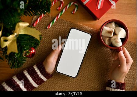 Une femme tenant une maquette d'écran blanc de smartphone et un chocolat chaud avec guimauves sur une table en bois avec décoration de Noël. vue de dessus Banque D'Images