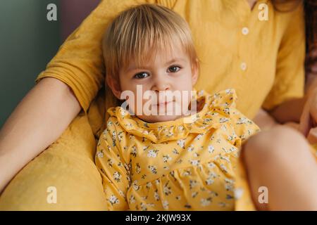 Petite fille drooling dans une robe jaune, s'appuyant sur la mère, se trouve sur le lit et regarde la caméra Banque D'Images