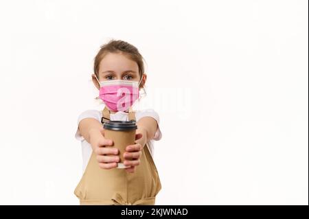 Jolie petite fille dans le tablier du chef et masque de sécurité médical rose, donnant sur l'appareil photo une boisson chaude à emporter dans une tasse en papier Banque D'Images