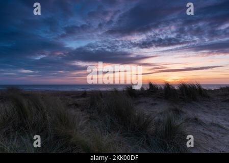 Un lever de soleil à l'aube avec des nuages sombres, à travers l'herbe de maram et les dunes à la plage de Winterton, à Norfolk. Winterton, février 2022 Banque D'Images