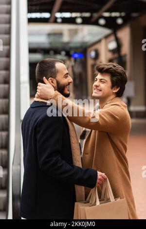 vue latérale de gai gai homme embrassant le cou de l'ami tendance tenant des sacs de shopping à l'extérieur, image de stock Banque D'Images