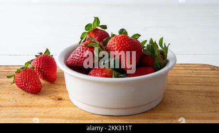 vue rapprochée d'un bol en céramique blanche avec des fraises rouges fraîches à l'intérieur et deux d'entre elles placées sur une table en bois Banque D'Images