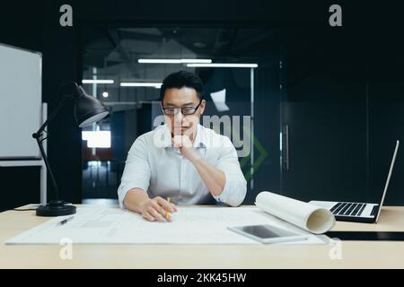 Un beau jeune homme, un architecte asiatique, concepteur, ingénieur, s'assoit à un bureau au bureau, à une table avec des carnets et des documents. Il se concentre sur la fabrication de dessins, le développement d'un plan. Banque D'Images