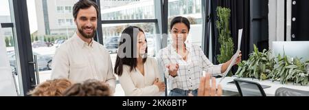 bonne femme multiraciale tenant des documents et pointant de la main près de collègues multiethniques d'affaires, bannière Banque D'Images