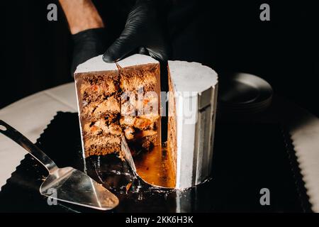Des gâteaux incroyables. Un chef aux gloved noirs coupe un gâteau de mariage au chocolat. Le gâteau de mariage est délicieux à l'intérieur sur un fond noir.Grand gâteau en blanc Banque D'Images