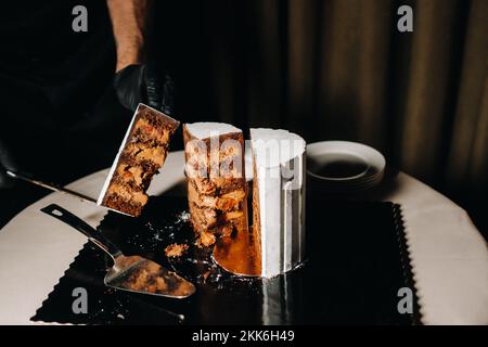 Des gâteaux incroyables. Un chef aux gloved noirs coupe un gâteau de mariage au chocolat. Le gâteau de mariage est délicieux à l'intérieur sur un fond noir.Grand gâteau en blanc Banque D'Images