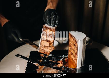 Des gâteaux incroyables. Un chef aux gloved noirs coupe un gâteau de mariage au chocolat. Le gâteau de mariage est délicieux à l'intérieur sur un fond noir.Grand gâteau en blanc Banque D'Images