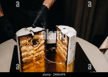 Des gâteaux incroyables. Un chef aux gloved noirs coupe un gâteau de mariage au chocolat. Le gâteau de mariage est délicieux à l'intérieur sur un fond noir.Grand gâteau en blanc Banque D'Images