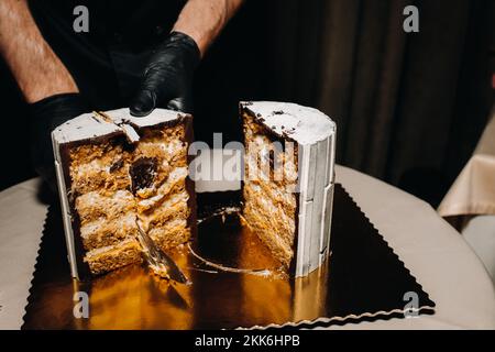 Des gâteaux incroyables. Un chef aux gloved noirs coupe un gâteau de mariage au chocolat. Le gâteau de mariage est délicieux à l'intérieur sur un fond noir.Grand gâteau en blanc Banque D'Images