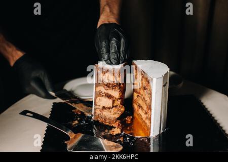 Des gâteaux incroyables. Un chef aux gloved noirs coupe un gâteau de mariage au chocolat. Le gâteau de mariage est délicieux à l'intérieur sur un fond noir.Grand gâteau en blanc Banque D'Images