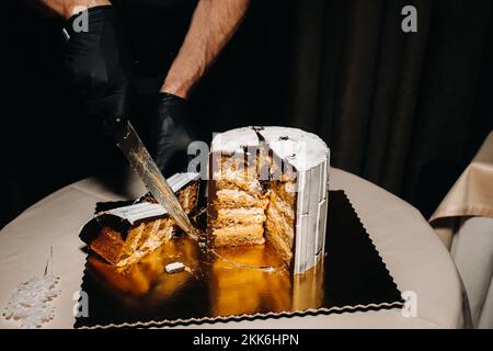 Des gâteaux incroyables. Un chef aux gloved noirs coupe un gâteau de mariage au chocolat. Le gâteau de mariage est délicieux à l'intérieur sur un fond noir.Grand gâteau en blanc Banque D'Images