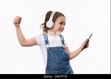 Adorable petite fille tenant le téléphone mobile dans ses mains, danse et écoute la musique sur son casque stéréo Banque D'Images