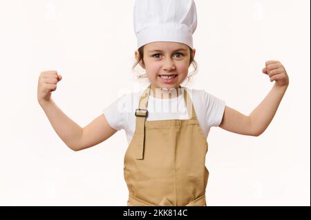 Bébé fille surjoyeuse vêtue comme confiseur dans le chapeau du chef et tablier clenches fisches, regarde l'appareil photo, sur fond blanc Banque D'Images