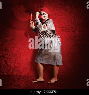 Scary clown doctor holding grande seringue médicale sur le point de donner une cpr le mur de ciment rouge. Médecine du mal Banque D'Images