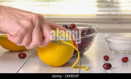 Femme mains peeling orange à l'aide de zester couteau. Recette de sauce à l'orange canneberge. Gros plan des ingrédients sur la table de cuisine Banque D'Images