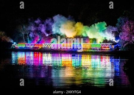 Kingswear, Devon, Royaume-Uni. 25th novembre 2022. Le train de lumière de Noël sur le chemin de fer à vapeur de Dartmouth, qui fonctionne entre le 25th novembre et le 30th décembre 2022, se reflète dans l'eau calme de la rivière Dart alors qu'elle se dirige vers Kingswear à Devon lors de son voyage au départ de Paignton. Les wagons et les locomotives à vapeur du service de fête sont éclairés à l'intérieur et à l'extérieur par des lumières colorées. Crédit photo : Graham Hunt/Alamy Live News Banque D'Images