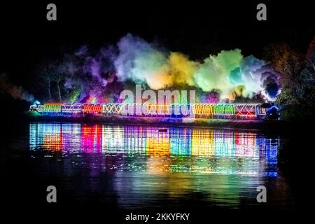 Kingswear, Devon, Royaume-Uni. 25th novembre 2022. Le train de lumière de Noël sur le chemin de fer à vapeur de Dartmouth, qui fonctionne entre le 25th novembre et le 30th décembre 2022, se reflète dans l'eau calme de la rivière Dart alors qu'elle se dirige vers Kingswear à Devon lors de son voyage au départ de Paignton. Les wagons et les locomotives à vapeur du service de fête sont éclairés à l'intérieur et à l'extérieur par des lumières colorées. Crédit photo : Graham Hunt/Alamy Live News Banque D'Images