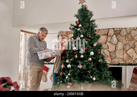 Couple de personnes âgées, Noël et décoration d'arbre ensemble en préparation pour la saison des fêtes à la maison. Homme et femme âgés qui pendent de la lumière ou de la décoration Banque D'Images