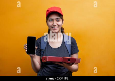 Jeune femme de livraison en uniforme noir et chapeau rouge tenant des boîtes à pizza montrant le téléphone mobile regardant devant sourire Banque D'Images