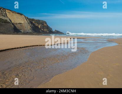 Vue sur la plage vide de Praia do Carvalhal avec un petit ruisseau menant à l'océan, des vagues, des falaises et des pierres, du sable doré humide et de la végétation verte à Rota sauvage Banque D'Images