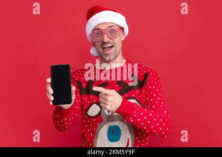 homme surpris en chandail et chapeau du père noël. un gars de noël pointant du doigt sur l'écran du téléphone Banque D'Images