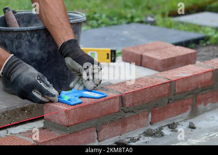 Construire un mur de briques rouges. Mains mâles tenant la truelle de brique et l'outil de mesure de niveau. Travaux de maçonnerie en détails. Photo de près du chantier de construction. Banque D'Images