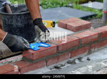 Construire un mur de briques rouges. Mains mâles tenant la truelle de brique et l'outil de mesure de niveau. Travaux de maçonnerie en détails. Photo de près du chantier de construction. Banque D'Images