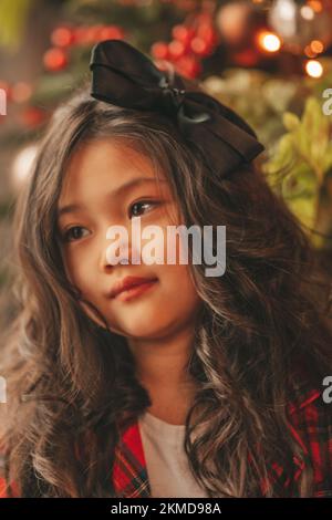 Bonne petite fille de rêve de japon regardant loin en attendant miracle de Santa posant près de l'arbre de noël. Asiatique Kid noir cheveux bouclés dans un costume rouge à carreaux fête Banque D'Images