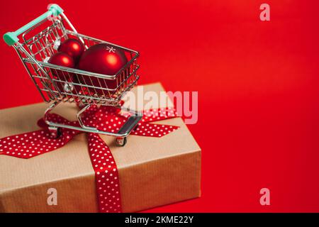 Vente de Noël bannière Web avec mini chariot avec boîte cadeau et décorations de Noël rouges boules sur fond rouge. Achats de Noël minimaux Banque D'Images