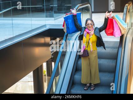 Une dame de premier plan célèbre avec des sacs de shopping dans les escaliers mécaniques du centre commercial. Femme descendant l'escalier roulant. Banque D'Images