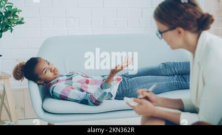 Une jeune fille afro-américaine déprimée parle au psychologue allongé sur un canapé et parle tandis que la femme thérapeute en lunettes écoute et écrit. Concept d'aide et de spécialistes. Banque D'Images