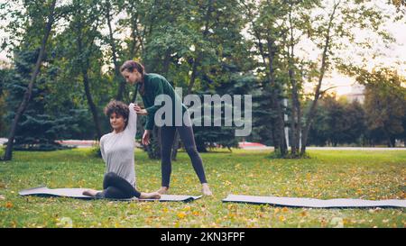 Diligent élève de yoga est d'apprendre la position de visage de vache assis sur le tapis et les bras d'étirement tandis que l'enseignant expérimenté l'aide, les filles parlent et rient à l'extérieur de ptactice. Banque D'Images