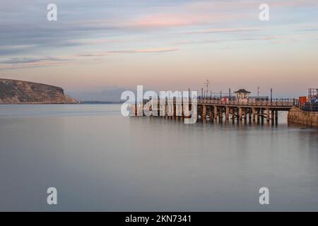 Une longue exposition claire en novembre soir regardant à travers les eaux lisses et vides le long de Swanage Pier avec un ciel rose Banque D'Images