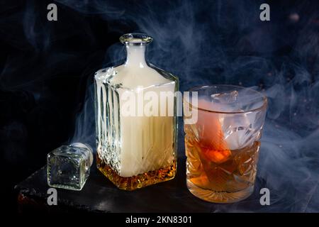 Couvert de verre de whisky fumé avec fumée et verre de whisky avec glace et zeste d'orange sur plaque d'ardoise. Mélange de whisky avec de la fumée pour donner un Banque D'Images