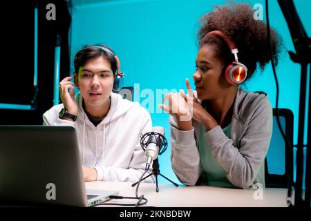 Jeune femme afro-américaine et homme asiatique un podcast interview et conversation ensemble en studio, homme et femme hôte portant casque parler avec Banque D'Images
