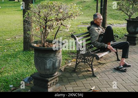 Un vieil homme du sud-est asiatique est assis sur la chaise publique près du pot dans le parc Banque D'Images